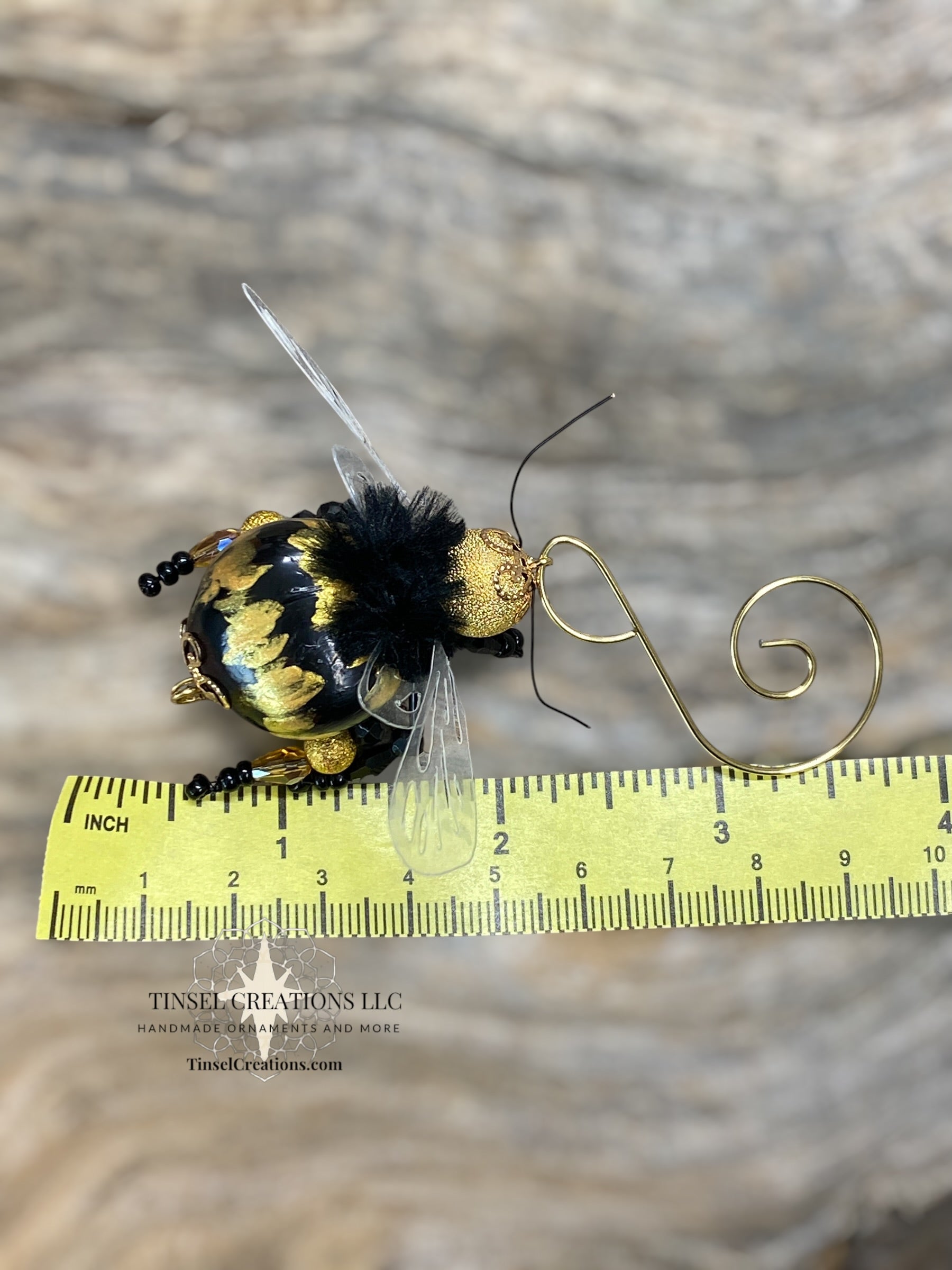 Decorative bee ornament with gold and black details on a wooden surface, measured against a ruler.