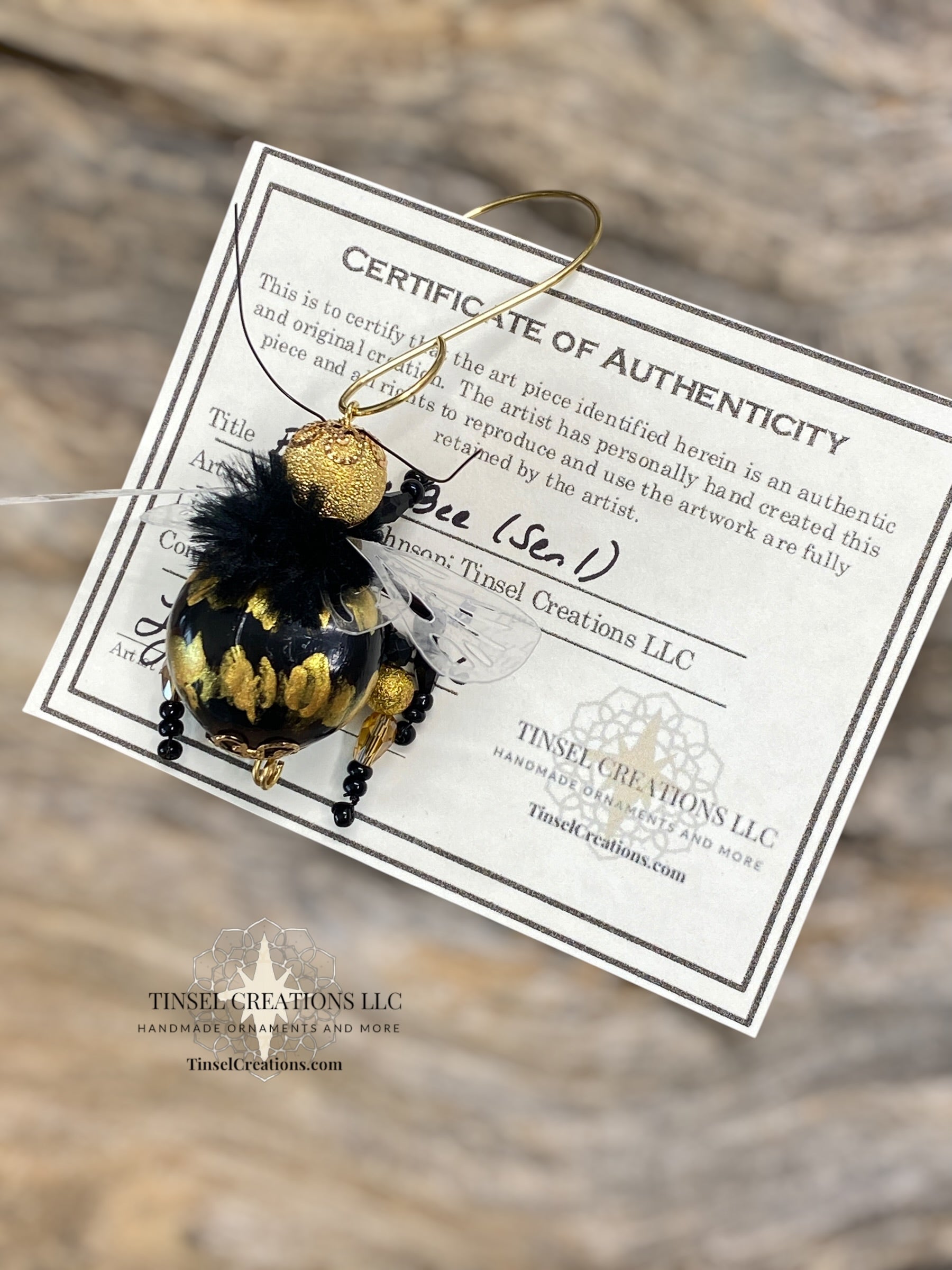 Decorative bee ornament with gold and black details on a Tinsel Creations LLC certificate of authenticity against a blurred background.