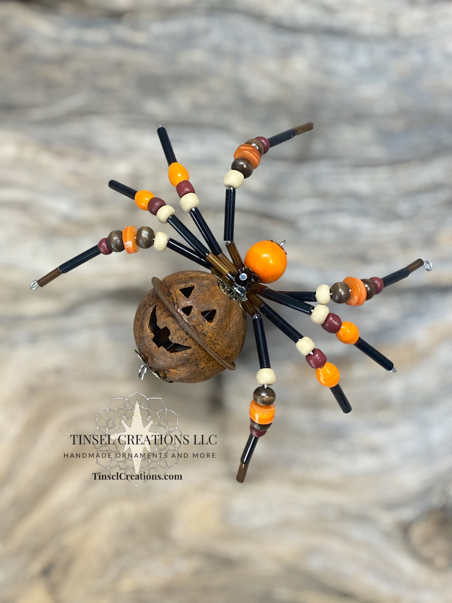 Decorative spider with beaded legs and a pumpkin head on a textured surface, featuring Tinsel Creations LLC branding.