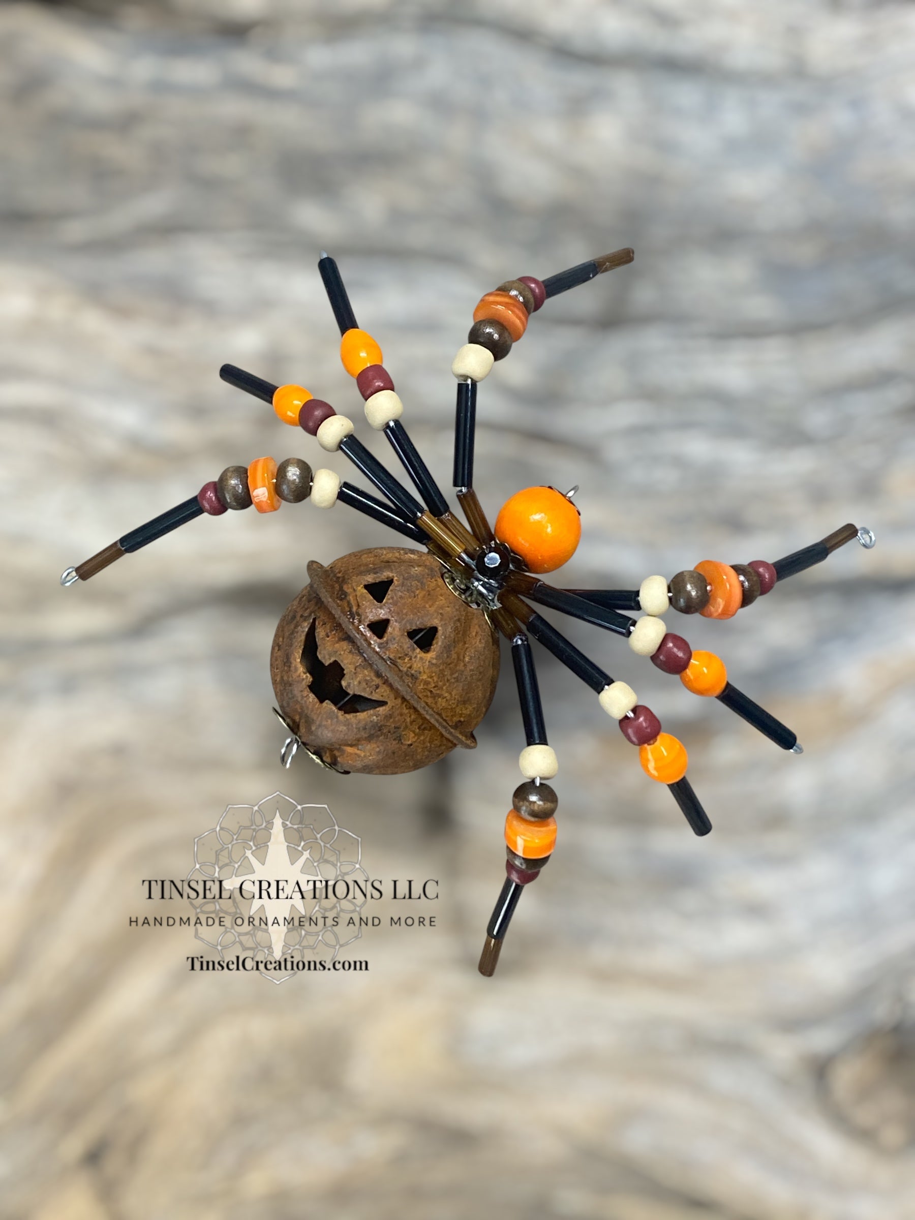 Decorative spider with beaded legs and a pumpkin head on a textured surface, featuring Tinsel Creations LLC branding.